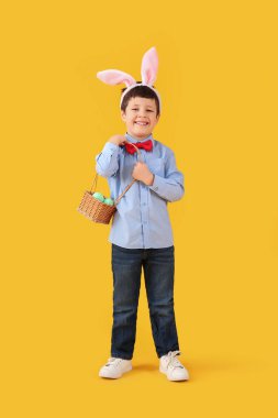 Cute little boy with bunny ears and Easter basket on yellow background