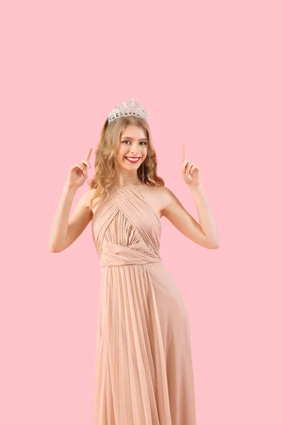 Teenage girl in tiara and prom dress pointing at something on pink background