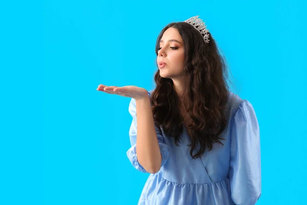 Teenage girl in tiara and prom dress blowing kiss on blue background