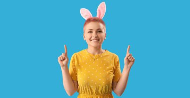 Young woman with Easter bunny ears pointing at something on blue background