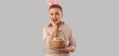 Surprised young woman with bunny ears and basket of Easter eggs on grey background