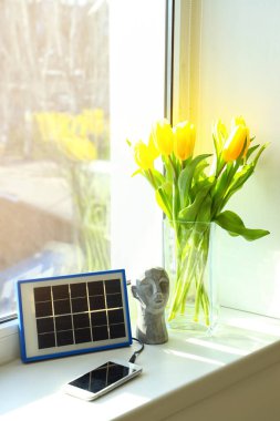 Portable solar panel charging mobile phone, decor and vase with tulips on windowsill in room