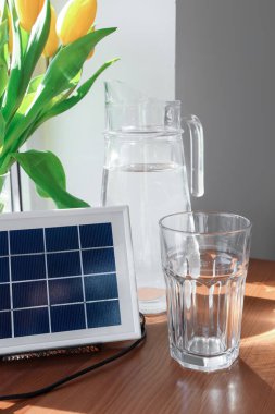 Portable solar panel, jug of water and vase with tulips on table near window, closeup