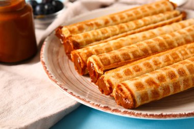 Plate of delicious wafer rolls with boiled condensed milk on blue background, closeup