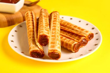 Plate of delicious wafer rolls with boiled condensed milk on yellow background