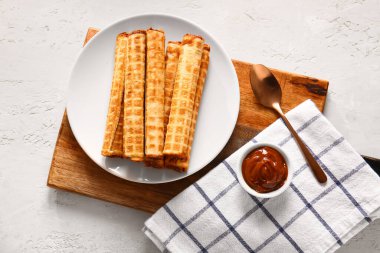 Plate with delicious wafer rolls and bowl of boiled condensed milk on white grunge background