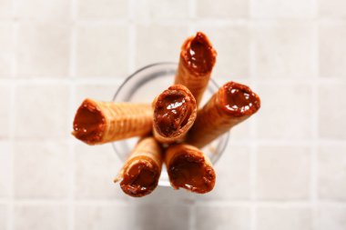 Jar of delicious wafer rolls with boiled condensed milk on tile background, closeup