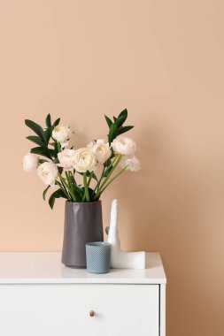 Vase with ranunculus flowers and decor on chest of drawers near beige wall