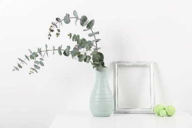 Blank frame, vase with eucalyptus branches and Easter eggs on white background