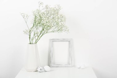 Blank frame, vase with gypsophila flowers and Easter eggs on white background