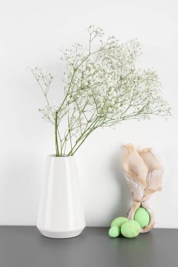 Vase with flowers and Easter eggs on table near white wall