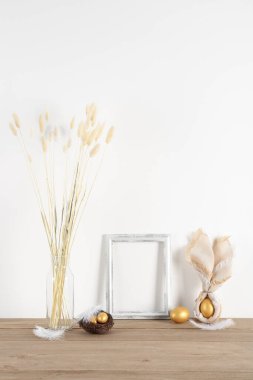 Blank frame, vase with dried flowers and Easter eggs on table near white wall