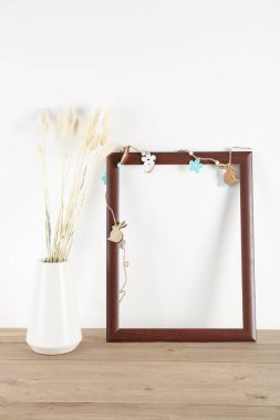 Blank frame with Easter decor and vase with dried flowers on table near white wall