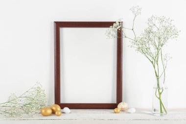 Blank frame, vase with flowers and Easter eggs on table near white wall