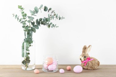 Vase with eucalyptus, Easter eggs and rabbit on table near white wall