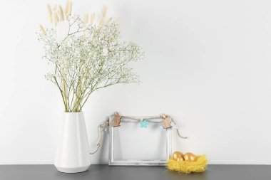 Blank frame, vase with flowers and Easter eggs on table near white wall