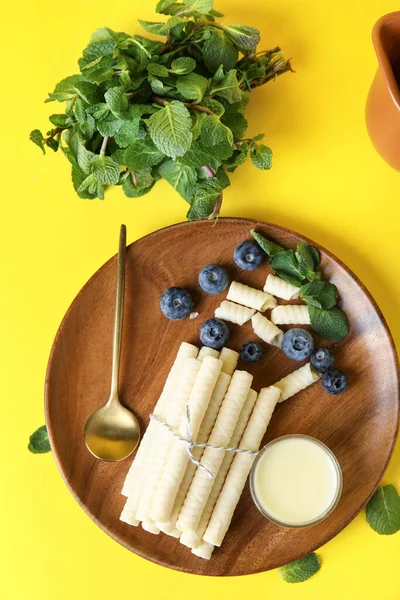 Board with delicious wafer rolls, condensed milk, blueberries and mint on yellow background