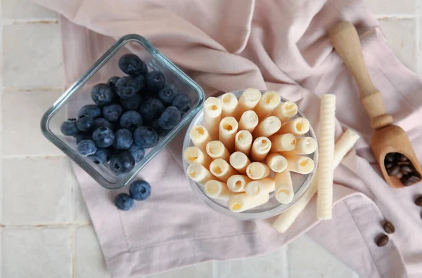 Glass of delicious wafer rolls and bowl with blueberries on tile table