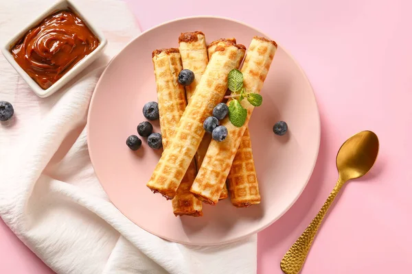 Plate of delicious wafer rolls with boiled condensed milk and blueberries on pink background