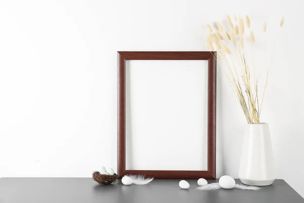 Blank frame, vase with dried flowers, Easter eggs and feathers on table near white wall