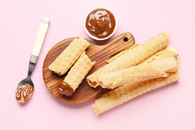 Board with delicious wafer rolls, spoon and boiled condensed milk on pink background