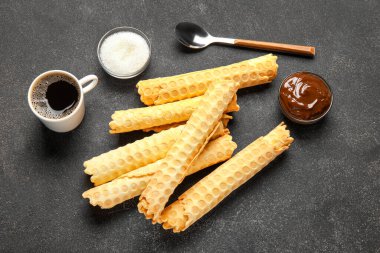 Delicious wafer rolls, boiled condensed milk, sugar and cup of coffee on black grunge background