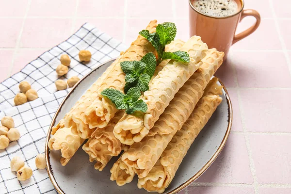 Plate with delicious wafer rolls, mint, hazelnuts and cup of coffee on pink tile background