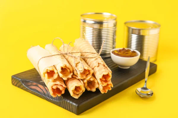 Board with stack of delicious wafer rolls on yellow background