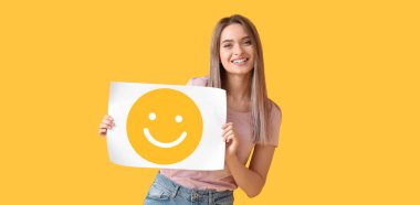 Pretty young woman holding paper with smiling emoticon on yellow background