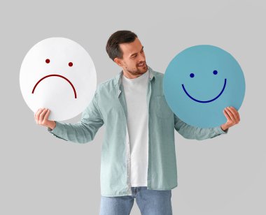 Handsome man holding happy and sad emoticons on light background 