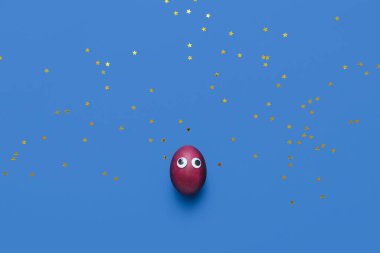 Funny Easter egg with googly eyes and glitter on blue background