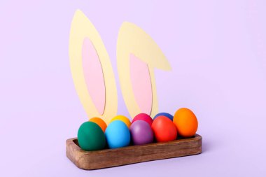 Box with Easter eggs and bunny ears on lilac background