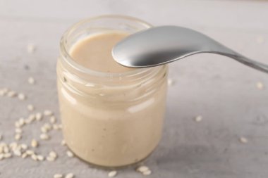 Glass jar of tasty tahini and spoon on light background, closeup