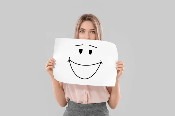 Pretty Young Woman Holding Paper Drawn Smiling Emoticon Light Background — Zdjęcie stockowe