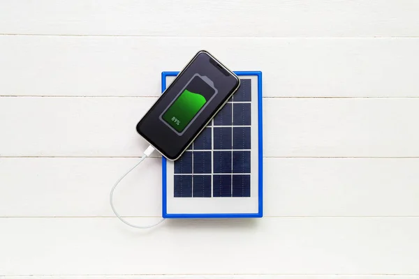 Portable solar panel charging mobile phone on white wooden background