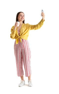 Akıllı telefonu ve kahvesi olan güzel genç bir kadın beyaz arka planda selfie çekiyor.