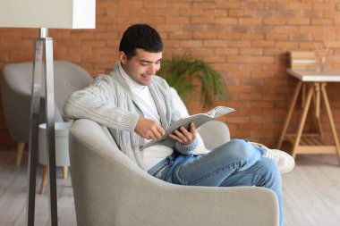 Genç adam evdeki gri koltukta dergi okuyor.
