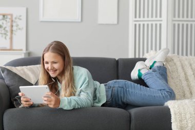 Kulaklıklı mutlu küçük kız evdeki kanepede tablet bilgisayar kullanıyor.