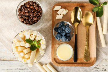 Glass of delicious wafer rolls and board with condensed milk on white wooden background