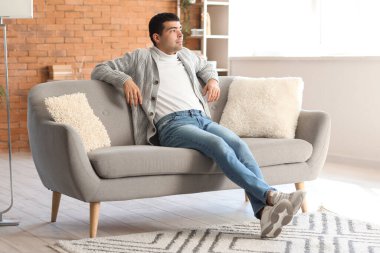 Genç adam evdeki gri kanepede dinleniyor.