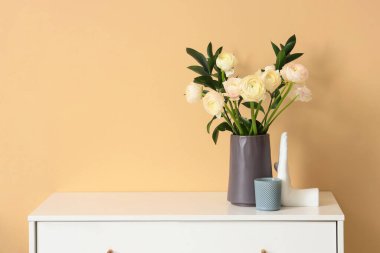 Vase with ranunculus flowers and decor on chest of drawers near beige wall