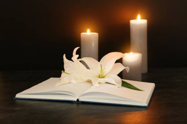Açık kitap, beyaz zambak çiçekleri ve koyu arkaplanda yanan mumlar.