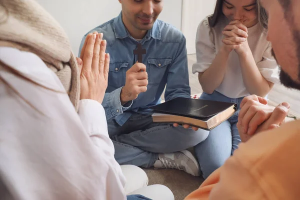 Group of people praying with Holy Bible and cross on floor at home