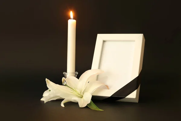 Blank funeral frame, white lily flowers and burning candle on dark background