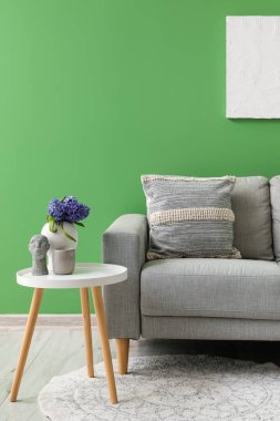 Yeşil duvarın yanındaki masada yastıklı ve vazo dolusu sümbül çiçekli gri kanepe.
