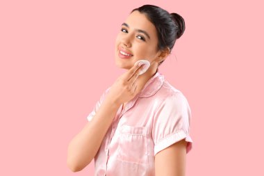 Genç bir kadın pembe arka planda pamuklu ped ile makyajı kaldırıyor.