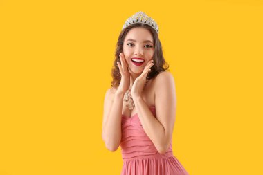 Beautiful surprised young woman in stylish prom dress and tiara on yellow background