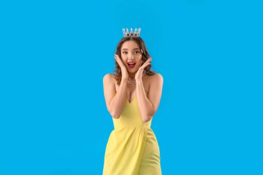 Mavi arka planda, balo elbisesi ve taç giymiş güzel, stresli genç bir kadın.