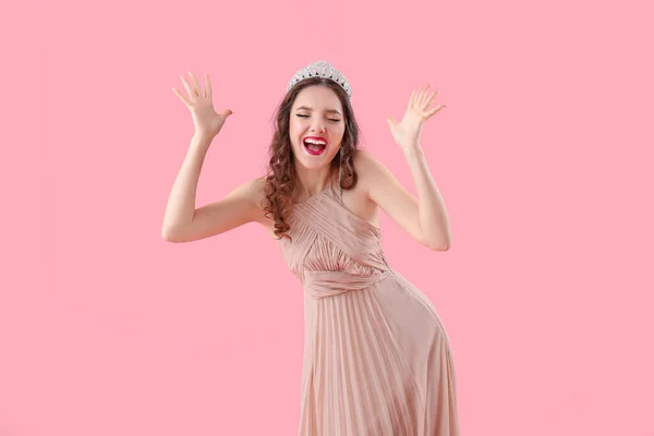 Stressed young woman in prom dress and tiara on pink background