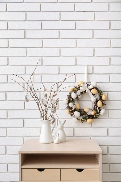 Table with willow branches in vase, toy rabbits and Easter wreath on white brick wall
